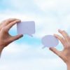 Close-up of hands holding speech bubbles, symbolizing communication and dialogue against a blue sky.
